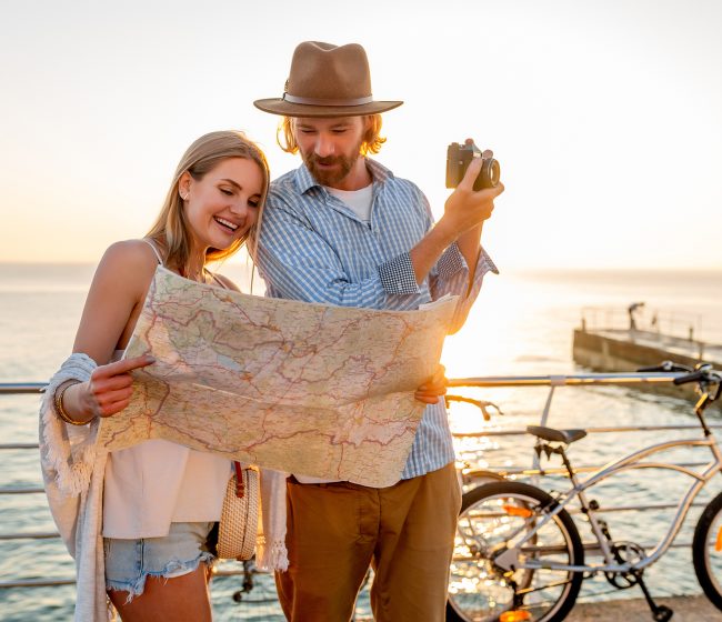 attractive happy couple traveling in summer on bicycles, man and woman with blond hair boho hipster style fashion having fun together, looking in map sightseeing taking photos on camera